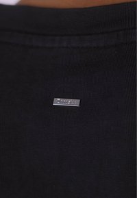 Sort bomulds T-shirt med en vævet tekstur og et lille sølv logo-mærke syet ind i stoffet nær kraven.