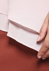 Lichtroze blouse stof met een transparante laag over een stevige roestkleurige kledingstuk. Gladde textuur, gebogen zoom en nette stikdetails.