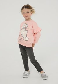 Różowa bluza z falbaniastymi rękawami z grafiką kota. W połączeniu z szarymi spodniami typu slim-fit i srebrnymi trampkami z białymi sznurówkami.