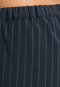 Mørkt stoff med hvite vertikale striper, med elastisk midje. Glatt tekstur, tett vevd materiale. Ingen tilleggsdetaljer.