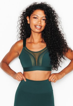 Donna con capelli lunghi e ricci che indossa un reggiseno sportivo verde scuro e leggings, in piedi con le mani sui fianchi su sfondo bianco.