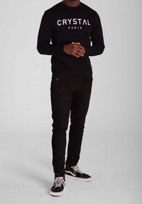 Sweatshirt en coton noir avec "CRYSTAL PARIS" imprimé en blanc, porté avec un jean noir et des baskets noires avec des accents et des lacets blancs.