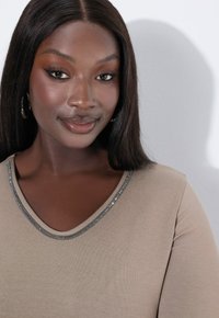Beige top s výstřihem do V, zdobený stříbrným flitrovým akcentem podél výstřihu. Hladká tkanina a dobře padnoucí střih jsou výrazně patrné.