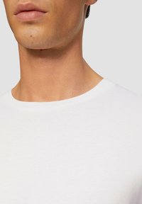 Witte katoenen t-shirt met een ronde halslijn, met een gladde textuur en minimale naden, van de schouder naar boven weergegeven.