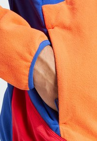 Fleec jaka s narančastim, plavim i crvenim dijelovima. Ima vidljivu džep i teksturirani materijal. Detaljna šavova duž šavova.