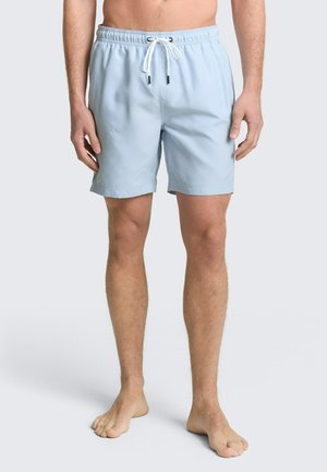 Mężczyzna w jasnoniebieskich kąpielówkach ze białym sznurowadłem, stojący boso na prostym jasnym tle.