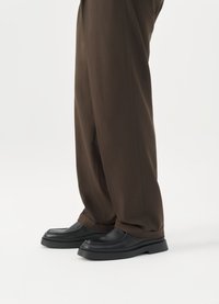 Svarta läderskor med en tjock, texturerad sula och rund tådesign, i kombination med löst sittande bruna byxor.