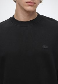 Jeune personne portant un sweat-shirt noir uni à col rond avec un petit logo Lacoste noir brodé sur la poitrine.