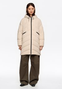 Abrigo acolchado beige con capucha, diseño acolchado diagonal, bolsillos con cremallera y cierre de cremallera negro. Combinado con pantalones sueltos de color verde oliva.