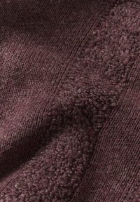 Nahaufnahme von strukturiertem, gestricktem Stoff in dunkelburgunderrot mit unterschiedlichen Stichmustern, die eine weiche, gemütliche Oberfläche erzeugen.