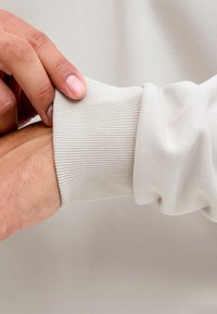 Kairė ranka reguliuoja šviesiai smėlio spalvos ilgomis rankovėmis megztos ribinės rankogalius ant dešinės riešo priešais vientisą foną.