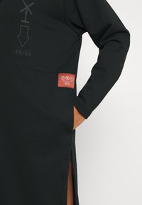 Vêtement noir au design épuré. Présente une fente sur le côté, des poignets côtelés et une étiquette rouge avec du texte. Texture douce, style décontracté.
