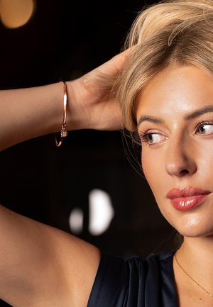 Rose gold bangle bracelet with a smooth, sleek design. The arm shown has a light skin tone and a delicate gold necklace. Dark background.