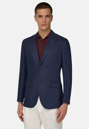 Man draagt een marineblauwe blazer over een bordeauxrood poloshirt en witte broek, staand tegen een effen lichte achtergrond.