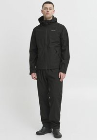 Mann iført svart hettejakke i vanntett materiale, matchende bukser og svarte slip-on sko, stående mot en enkel hvit bakgrunn.