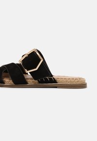 Sandale à glissière en suède noir avec une grande boucle rectangulaire en or, semelle en jute tressé et un design plat avec des brides croisées.