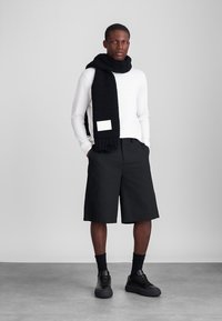 Must mustriga sall, mis on asetatud valge pika varrukaga särgi peale, koos mustade põlvikute pikkuses lühikeste pükste ja mustade jooksutossudega. Minimalistlik disain ja tekstuurilised detailid.