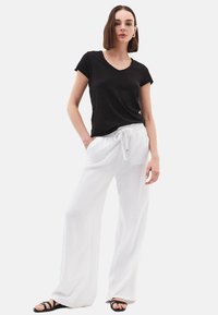 T-shirt nero con scollo a V abbinato a pantaloni bianchi in lino dalla vestibilità comoda con vita elasticizzata. Il modello indossa sandali neri con lacci.