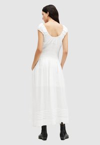 Robe maxi blanche sans manches avec un corsage froncé et un décolleté à volants, présentant des rayures verticales à l'ourlet ; portée avec des bottines noires.