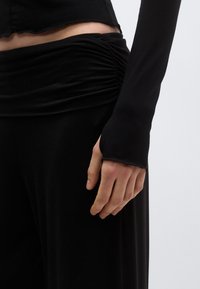 Μαύρο μακρυμάνικο τοπ με εφαρμοστή σχεδίαση και ραμμένο στη μέση με ρυτιδωτή ζώνη. Το ύφασμα φαίνεται ομαλό με λεπτή υφή και λεπτομέρειες με ακατέργαστες άκρες στα μανίκια.