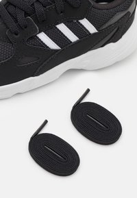 Sneaker nera con strisce bianche e suola bianca spessa, accanto a un paio di lacci neri arrotolati su una superficie bianca.