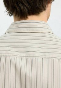 Camicetta beige chiaro con righe nere verticali, dotata di colletto classico e una texture liscia, leggermente lucida.