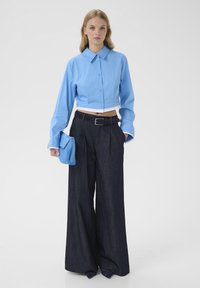 Chemise boutonnée bleu clair courte avec manches longues, associée à un pantalon large en denim foncé et une pochette bleue. Texture lisse, coupe ajustée.