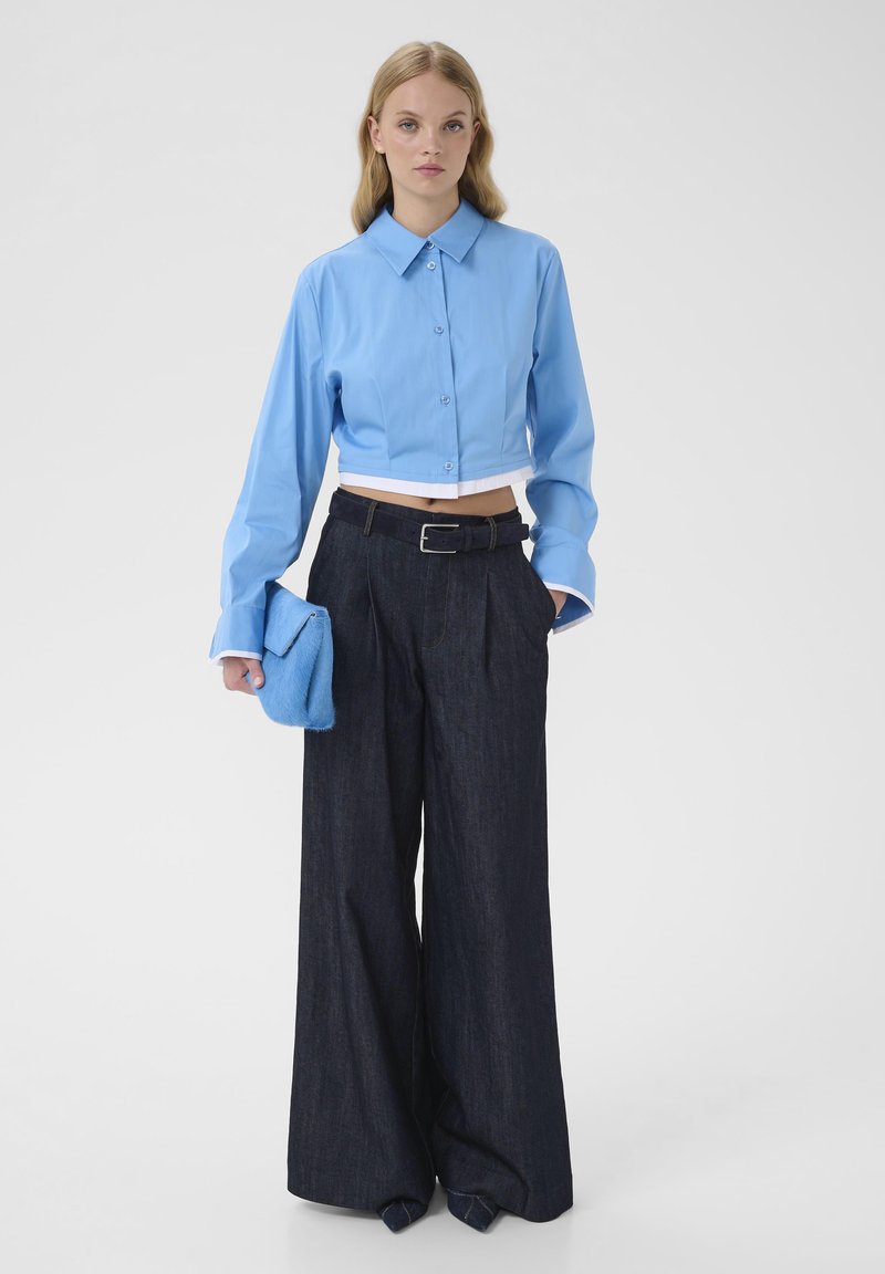 Chemise boutonnée bleu clair courte avec manches longues, associée à un pantalon large en denim foncé et une pochette bleue. Texture lisse, coupe ajustée.