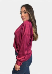 Top avvolgente in satin di un profondo magenta con maniche lunghe e vestibilità ampia, abbinato a jeans in denim a vita alta. Tessuto morbido e caduta fluida.