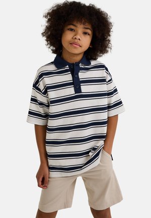 Polo rayé blanc avec des rayures horizontales bleu marine, doté de manches courtes, d'un col et d'une patte de boutonnage à trois boutons. Associé à un short beige.