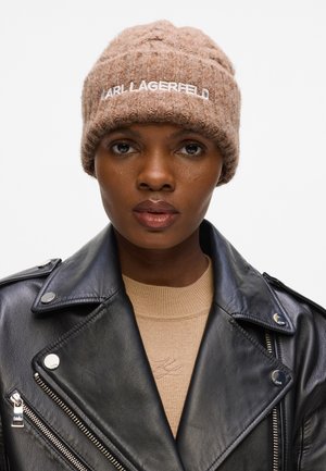 Bruine, gestructureerde beanie met het "Karl Lagerfeld" logo in het wit geborduurd, gedragen met een zwarte leren jas en een beige trui.