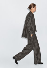 Vrouw die in een oversized donkergestreept pak loopt met handen in de zakken, draag een bril en heeft het haar opgestoken, tegen een effen lichte achtergrond.