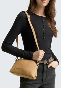 Cognac faux leren handtas met een gebogen vorm en een geperst logo. Het heeft een lange schouderband en is gecombineerd met een zwart geribbeld lange mouwen top.