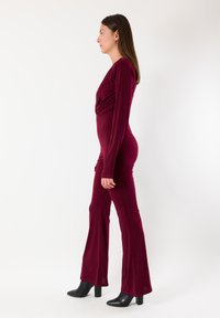 Tuta bordeaux a maniche lunghe con vita aderente, gambe svasate, realizzata in tessuto liscio. Abbinata a stivaletti neri con tacco a blocco.