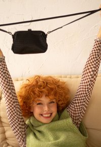 Černá texturovaná crossbody taška zavě�šená na popruhu, kterou drží osoba v zeleném pleteném topu a kostičkované košili s dlouhým rukávem.