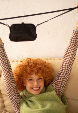 Borsa a tracolla nera con texture, sospesa su una tracolla, tenuta da una persona che indossa un top in maglia verde e una camicia a maniche lunghe a quadri.
