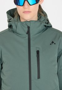 Grüne Softshell-Jacke mit hohem Kragen, schwarzen Reißverschlüssen und einer Brusttasche. Verfügt über ein dreieckiges Logo und eine verstellbare Kapuze mit Zugband.