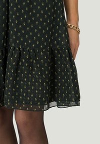 Jupe à motif vert foncé avec un ourlet à volants portée avec des collants noirs transparents et un bracelet en maillons dorés au poignet.