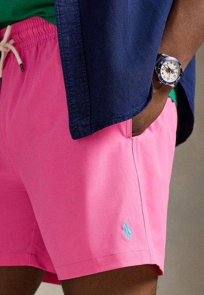 Ροζ μαγιό βερμούδα με ελαστική μέση και λευκό κορδόνι, με απλικέ λογότυπο σε γαλάζιο χρώμα. Φοριέται με μπλε σκούρο πουκάμισο και ρολόι.