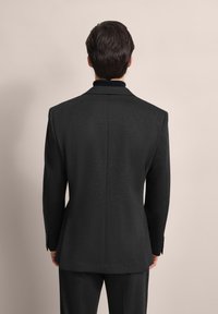 Schwarzer Blazer aus strukturiertem Stoff, mit taillierter Passform, Reverskragen und knopfbaren Manschetten, von hinten vor einem neutralen Hintergrund gezeigt.