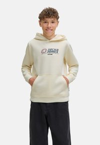 Un garçon souriant portant un sweat à capuche crème avec le logo "Jack & Jones", les mains dans les poches, se tenant devant un fond blanc uni.