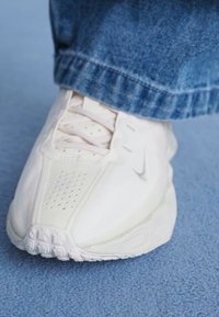 Białe sportowe buty z gładką skórzaną cholewką, perforacjami dla wentylacji, beżowymi sznurowadłami i fakturowaną podeszwą. Dżinsy częściowo widoczne.