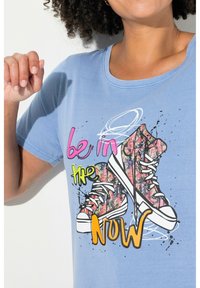 Camiseta de algodón azul con cuello redondo y mangas cortas, que presenta un gráfico de zapatillas coloridas y texto: "está en el AHORA."