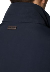 Marynarski kołnierz kurtki z teksturowanej tkaniny, z brązową skórzaną metką z wytłoczonym tekstem. Czyste linie i dopasowany krój.