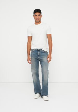 Valge lühikeste varrukatega t-särk väikese logoga, koos helesiniste laienevate teksadega, mille all on valged tennised. Üksildane taust.