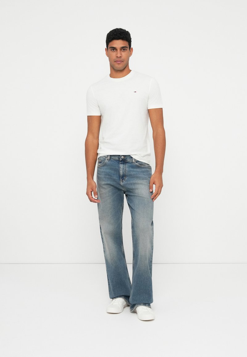 Λευκό T-shirt με κοντά μανίκια και μικρό λογότυπο, συνδυασμένο με ανοιχτόχρωμο τζιν με καμπάνα, φορώντας λευκά αθλητικά παπούτσια. Απλή φόντο.