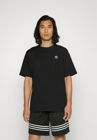 Čierne bavlnené tričko s okrúhlym výstrihom a krátkymi rukávmi; na ľavej strane hrudníka je malé biele logo Adidas; v kombinácii so vzorovanými čiernymi šortkami.