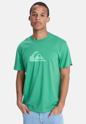 Junger Mann mit kurzen Haaren, trägt ein grünes T-Shirt mit einem Wellen- und Berglogo, Hände in den Taschen der hellblauen Jeans.