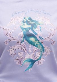 Modrá a zelená morská panna s vlajúci vlasmi a trblietavým chvostom s mierkami, obklopená dekoračnými špirálami a textom „Versace Jeans Couture“ na levanduľovej látke.