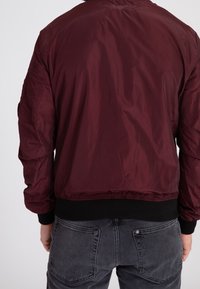 Giacca bomber color bordeaux con una texture liscia e lucida, polsini e vita elasticizzati; presenta una piccola toppa sul maniche sinistro.